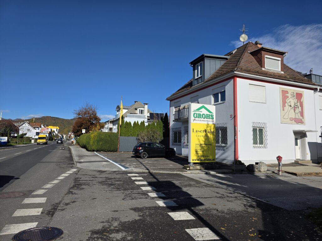 Straße mit Wohnhäusern und einem Werbeschild für LaserLabs Haarentfernung Mariatrost unter klarem blauem Himmel.