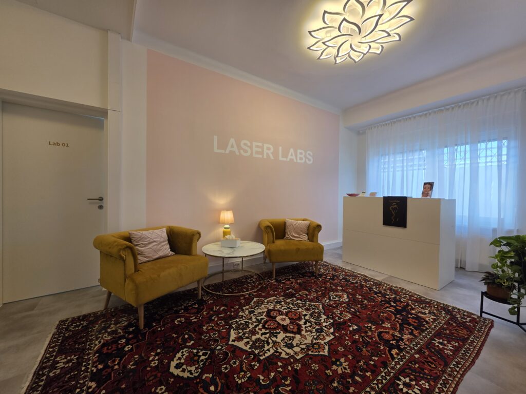 Modern eingerichteter Wartebereich mit zwei gelben Sesseln, einem kleinen Tisch, einem Teppich und einem Empfangstresen bei LaserLabs Haarentfernung Kalsdorf bei Graz.