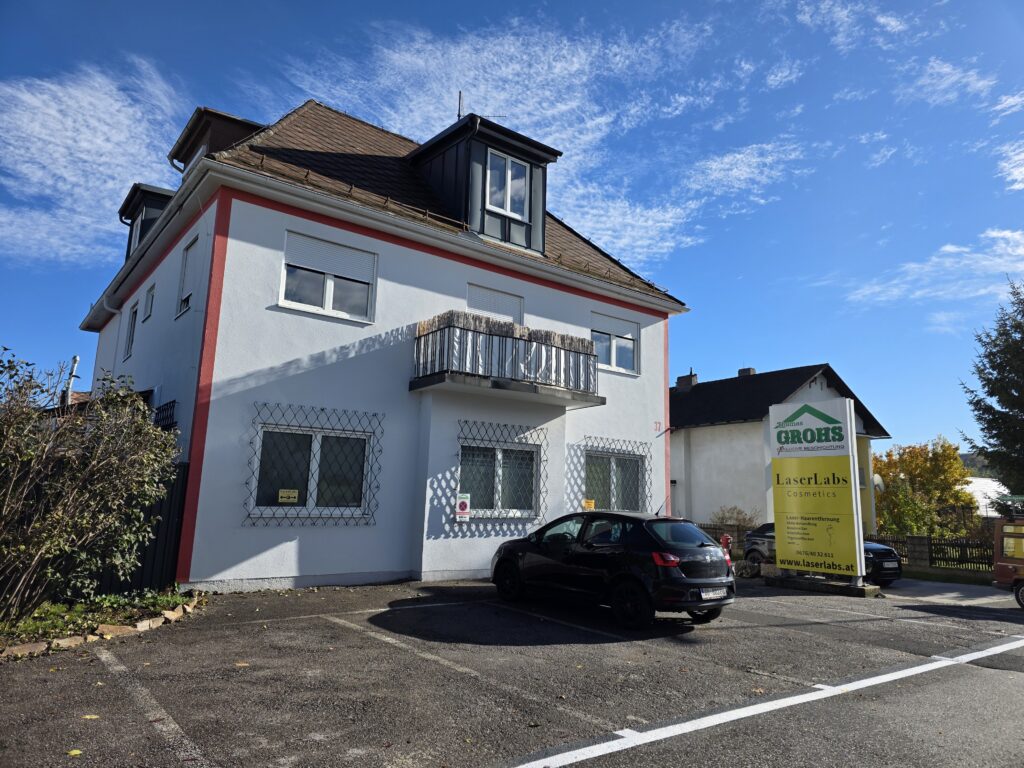 Modernes Gebäude mit Parkplatz und Werbetafel von LaserLabs Cosmetics, spezialisiert auf LaserLabs Haarentfernung Lannach, bei sonnigem Wetter und blauem Himmel.